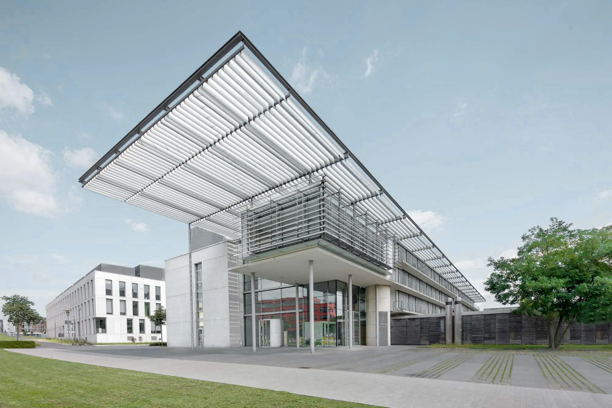 Gläsernes Vordach vor einem hellen Gebäude, getragen von schlanken Stützen